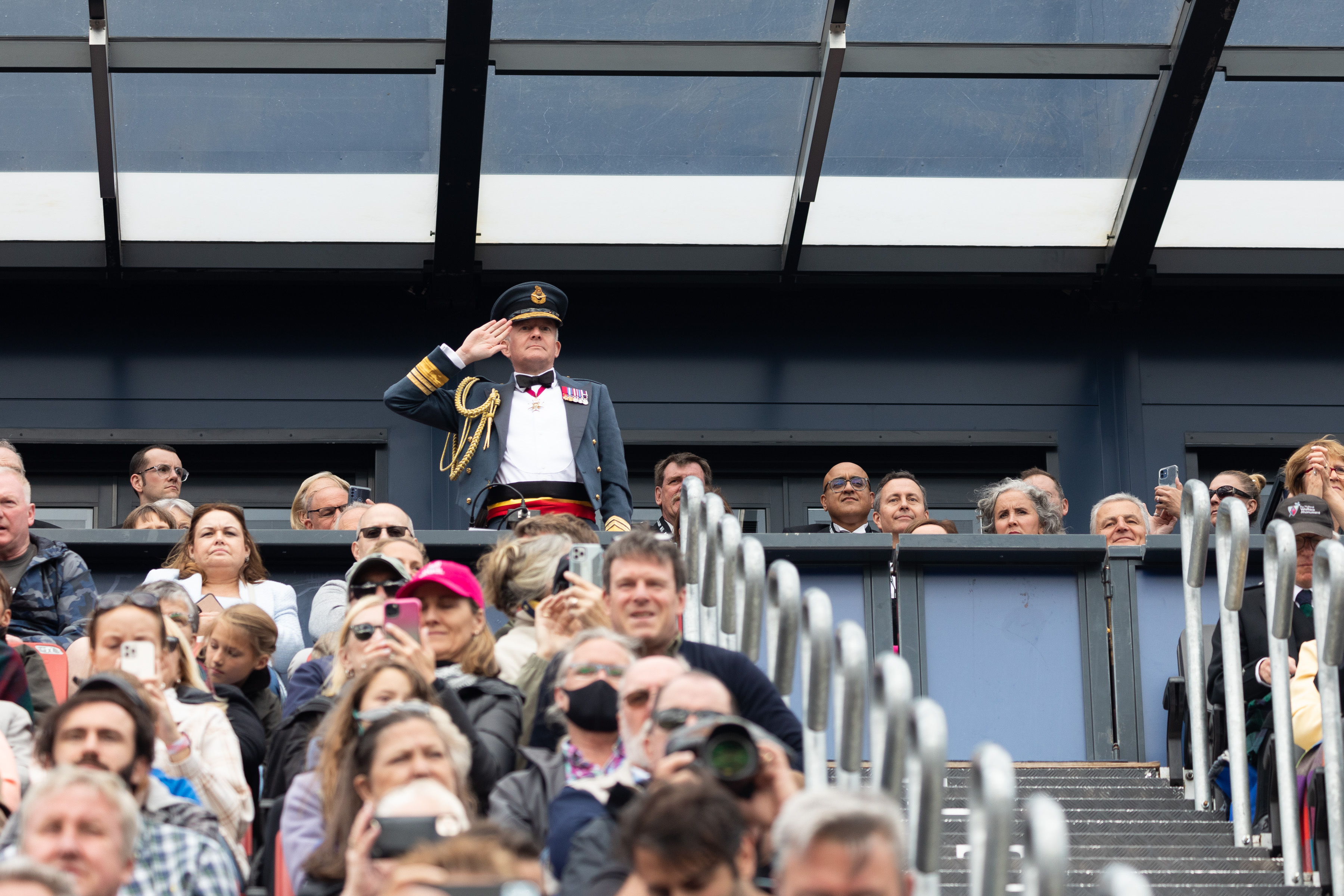 Image shows RAF aviator saluting from seating area amid public crowd.